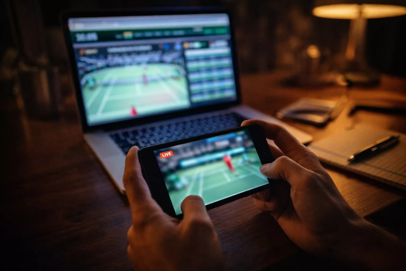 Person beobachtet ein Tennismatch live auf einem Smartphone neben einem Laptop